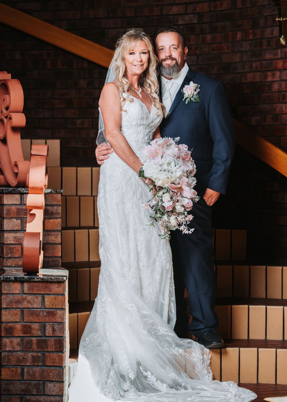 Newlywed couple standing on staircase against brick wall with bride holding pastel pink and white bouquet.