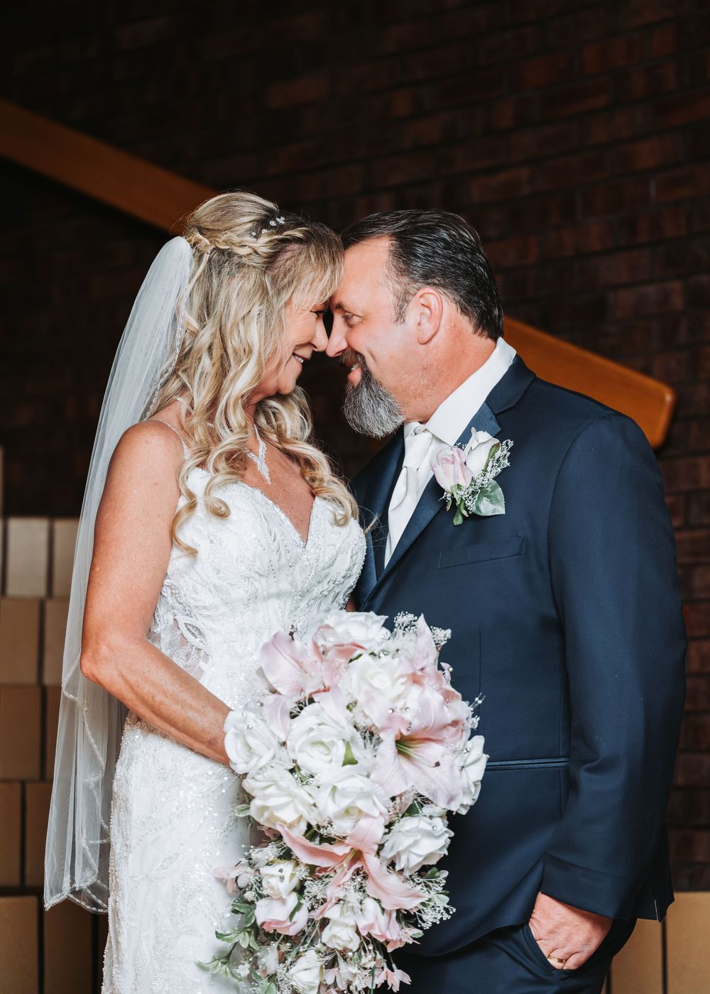 Elegant wedding couple shares intimate moment against brick wall while holding a soft pink and white peony bouquet.