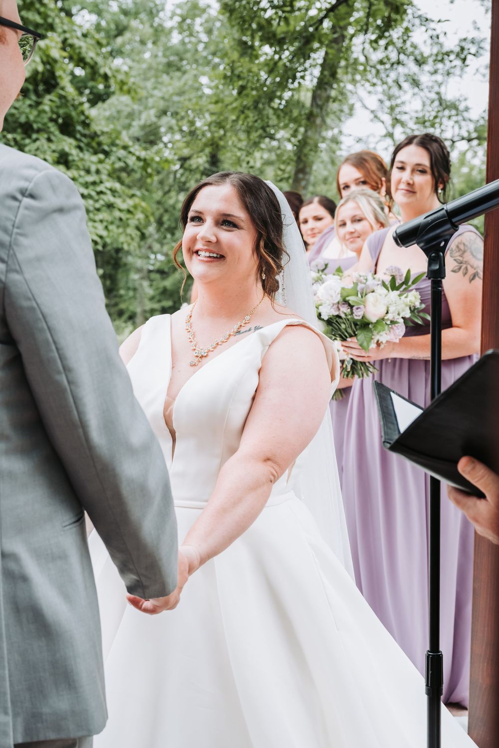 Wedding ceremony in outdoor garden setting with bride in white dress and bridesmaids wearing light purple gowns.
