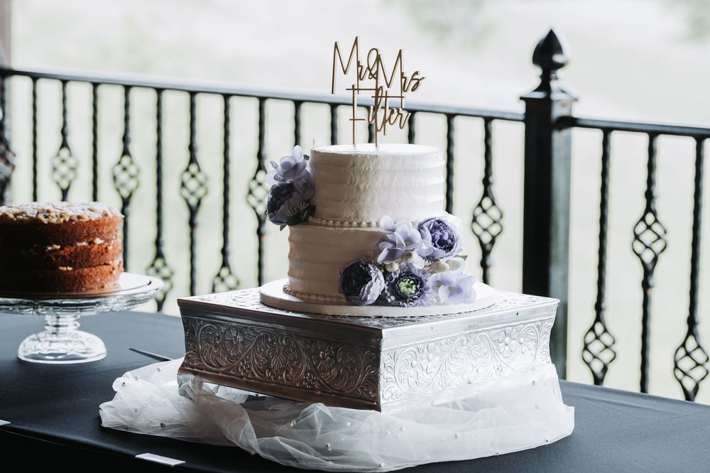 A two-tiered white wedding cake decorated with purple flowers sits on a silver stand next to an ornate railing outdoors.