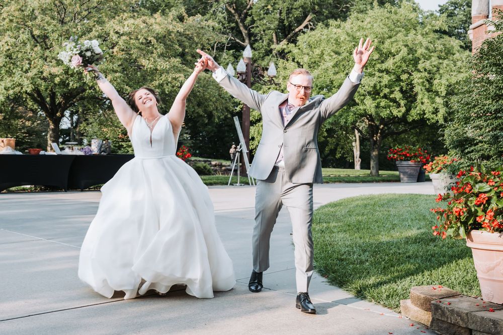 A couple in wedding attire jumps with joy on a garden pathway surrounded by lush greenery.