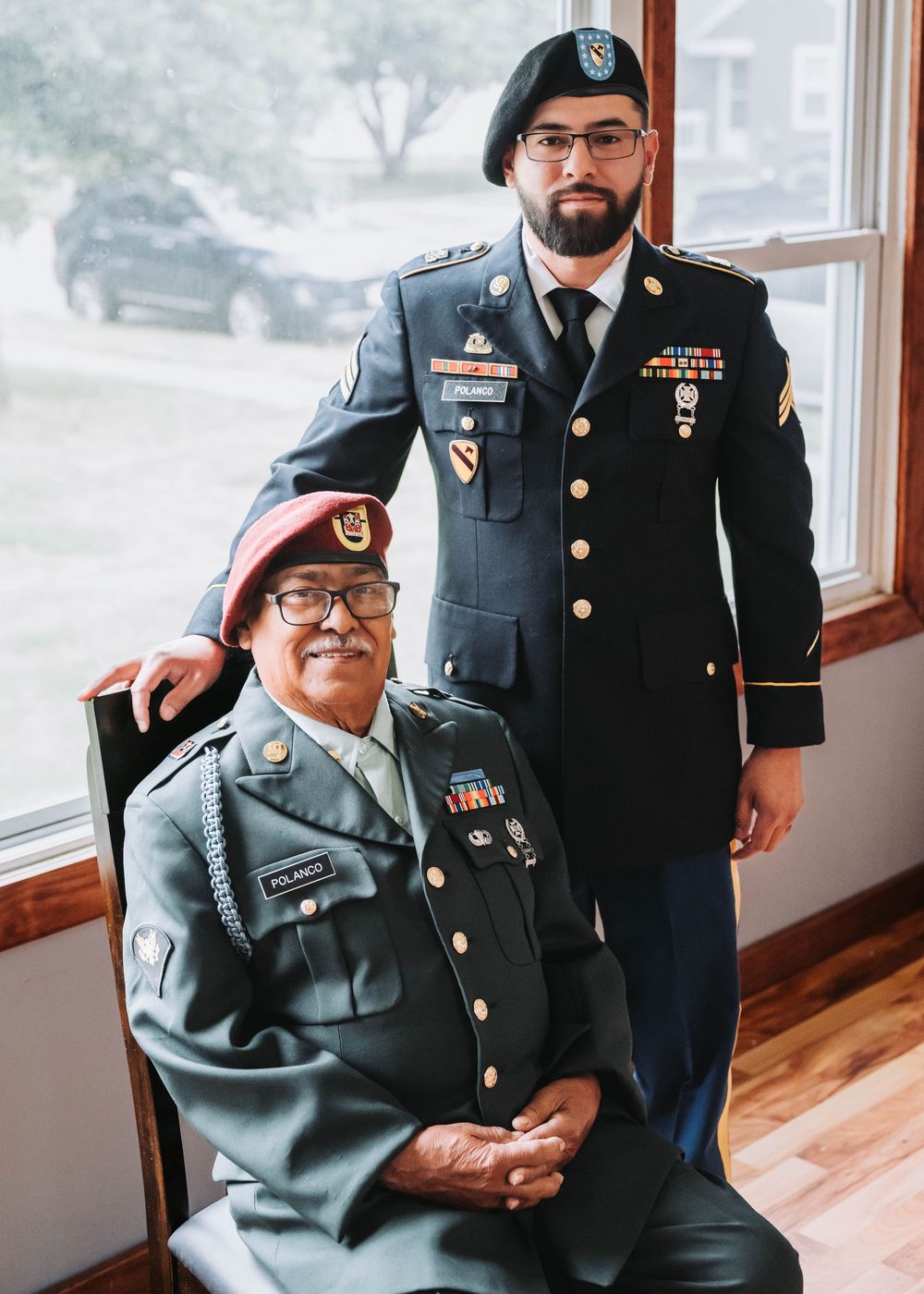 Two servicemembers in U.S. Army dress uniforms pose together by a window, showing military medals and decorations.