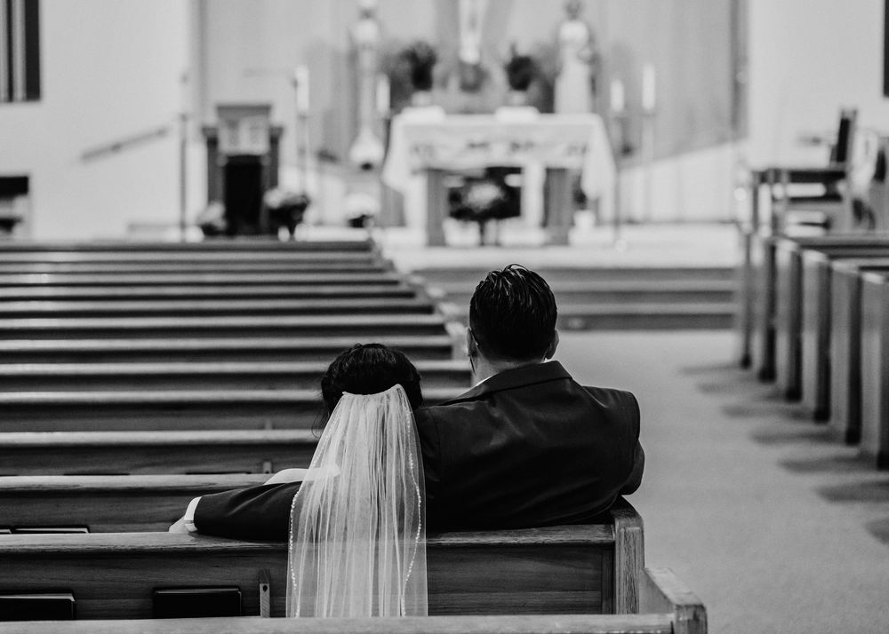 Two rows of wooden church pews are shown in black and white with a person in a veil walking down the aisle.