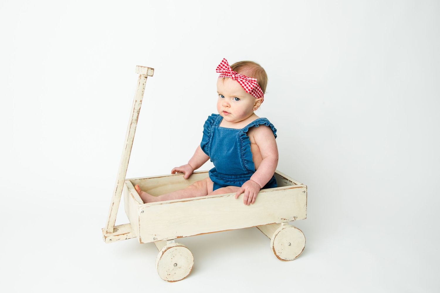 Baby wearing blue dress and pink headband sits in vintage white wooden wagon against white background.