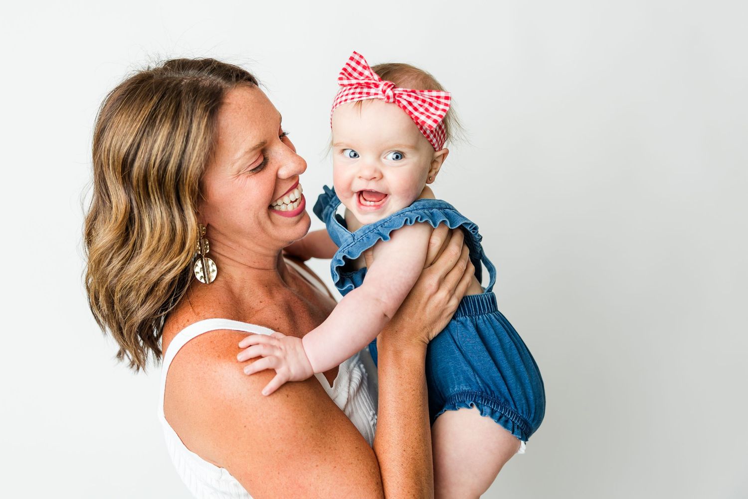 A joyful moment shared between parent and baby wearing a blue romper and red bow headband against a white background.
