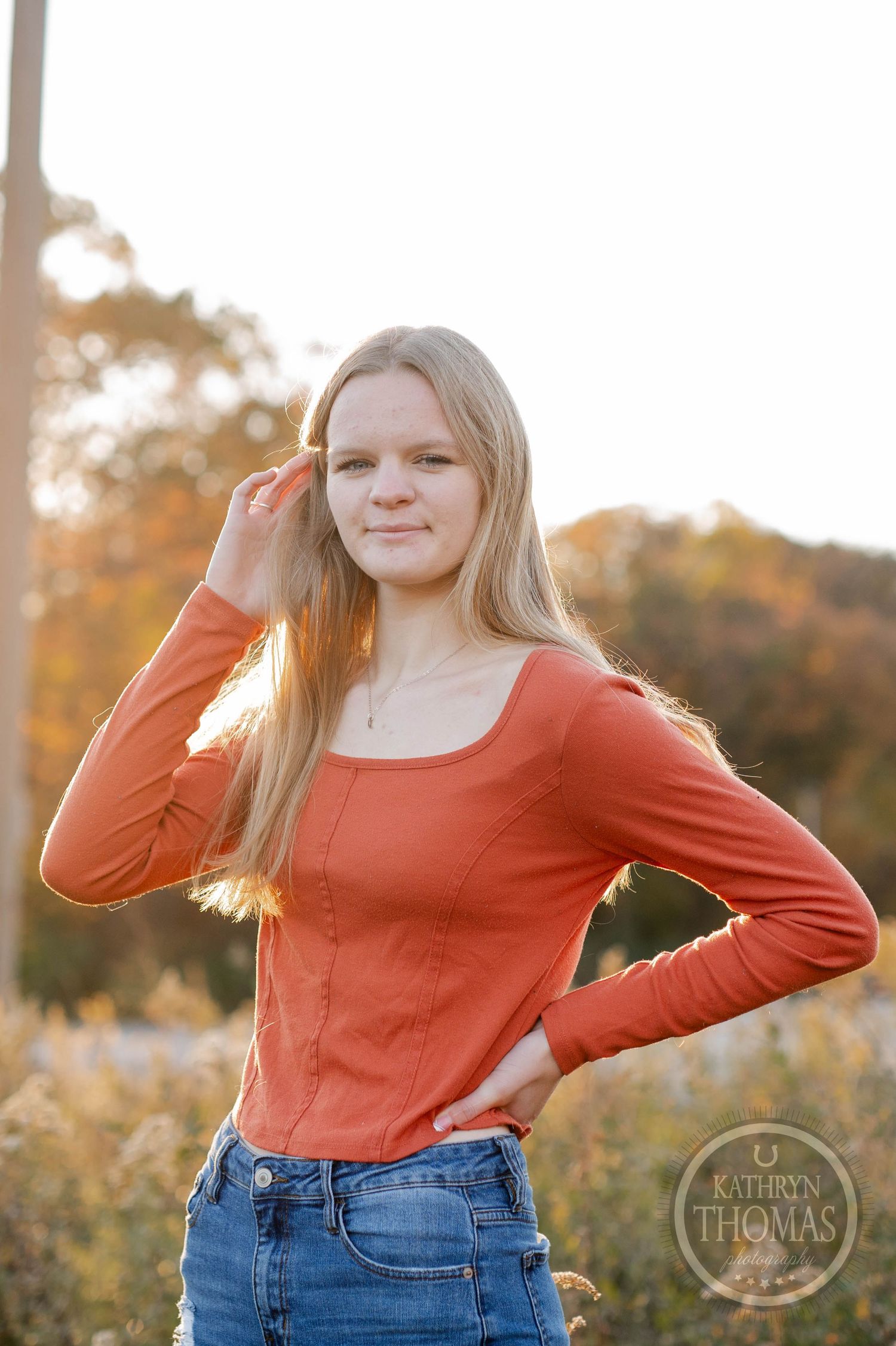 Person in coral long-sleeve top and jeans poses outdoors with autumn trees in background.
