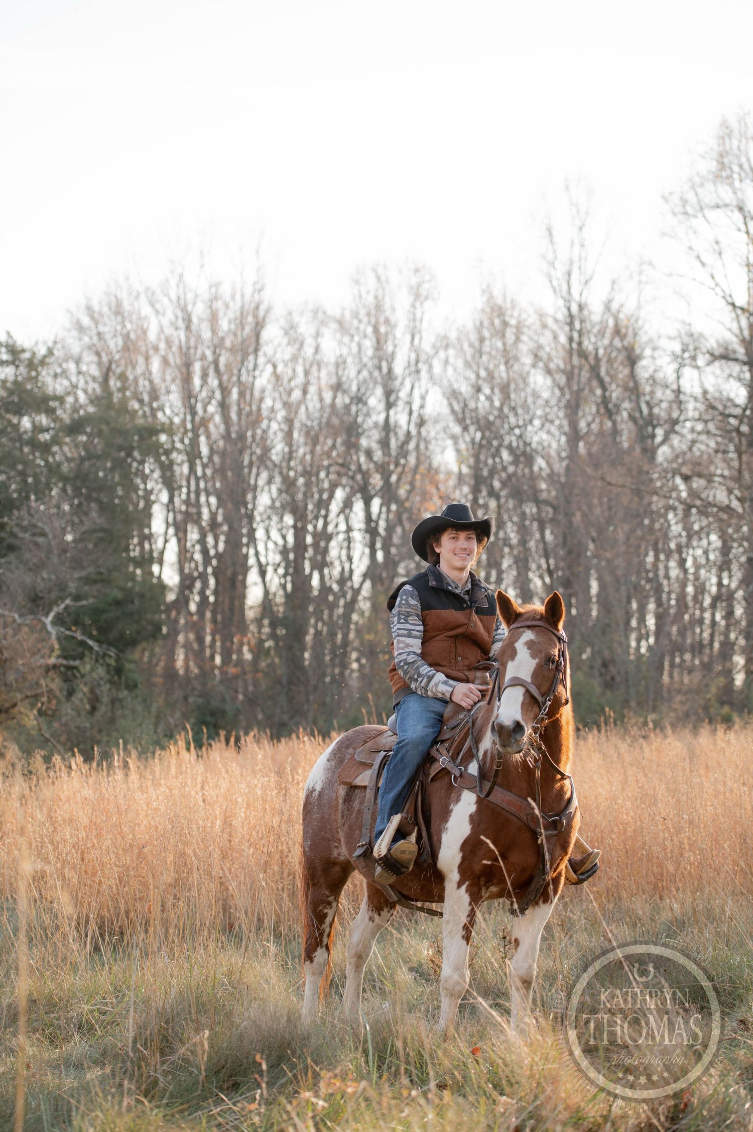 A paint horse and rider stand in a field of tall grass during sunset with bare winter trees behind.