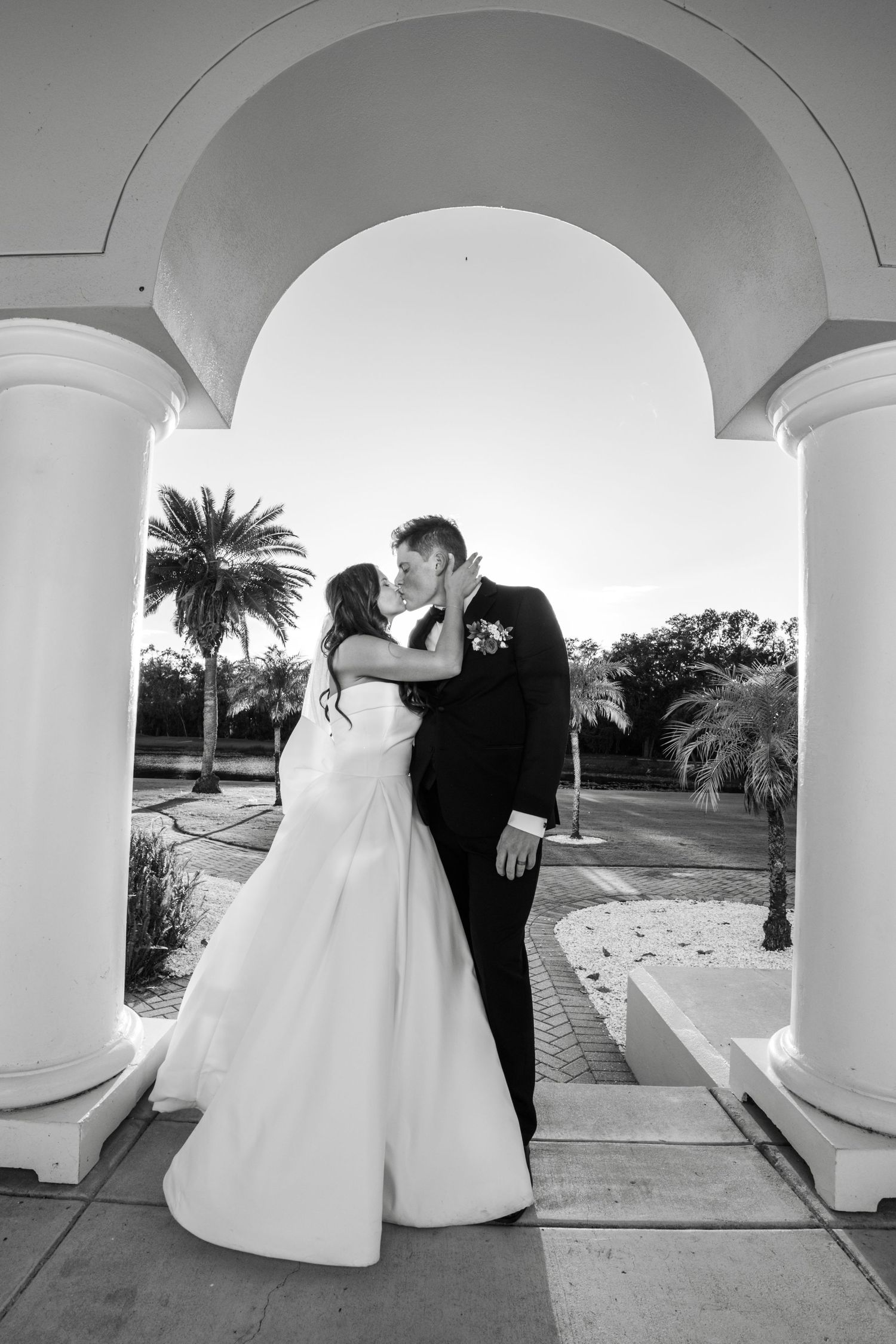 Romantic black and white wedding photo showing couple kissing under elegant white archway with palm trees in background.