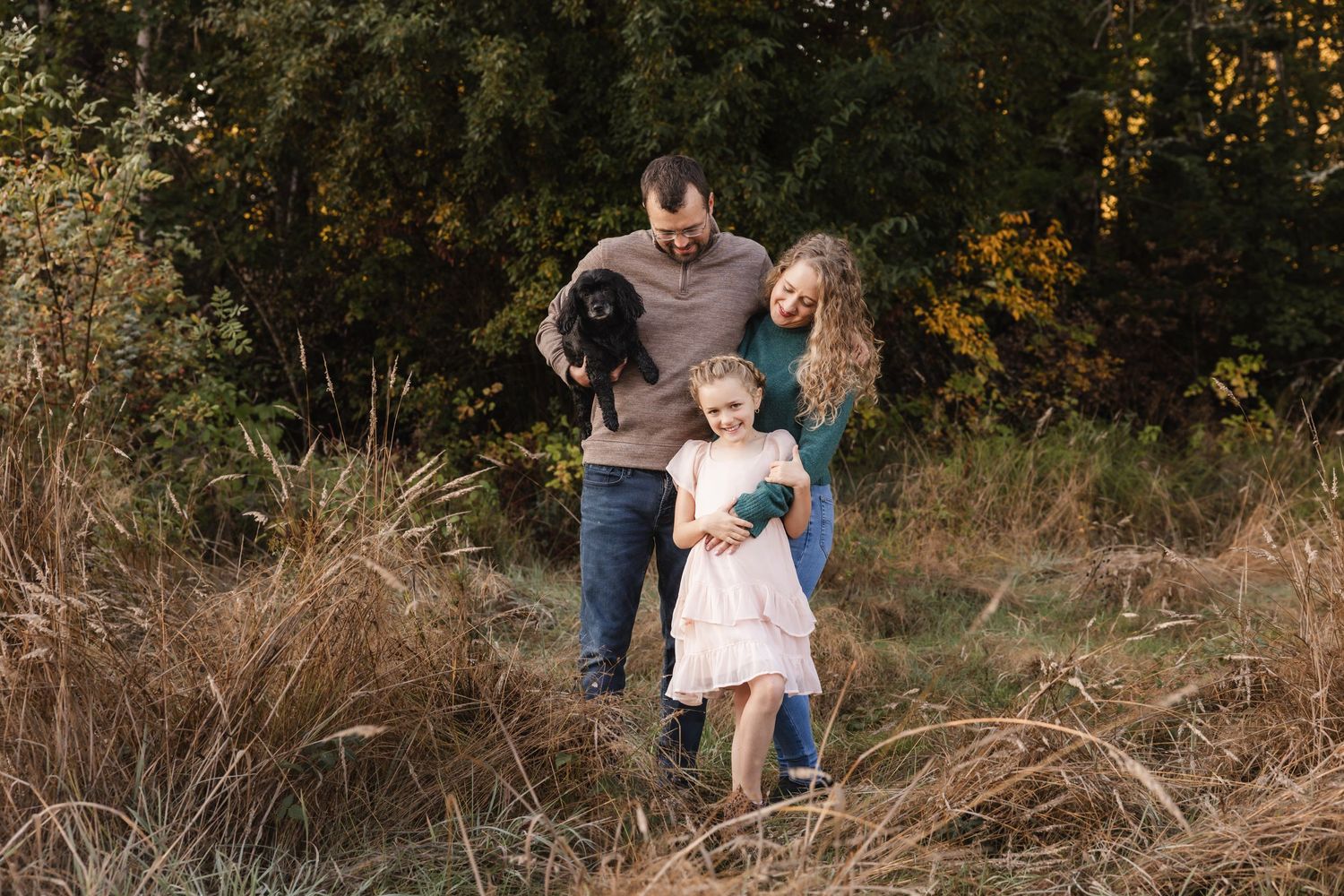 A family poses together in a rustic outdoor setting amid tall grass and autumn foliage at sunset.