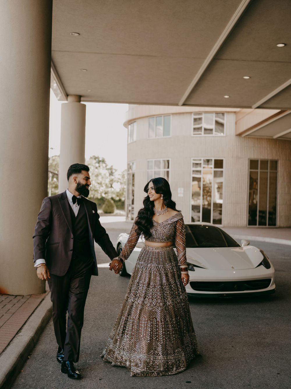 A couple in elegant attire walk hand in hand near a white luxury car outside a modern building.