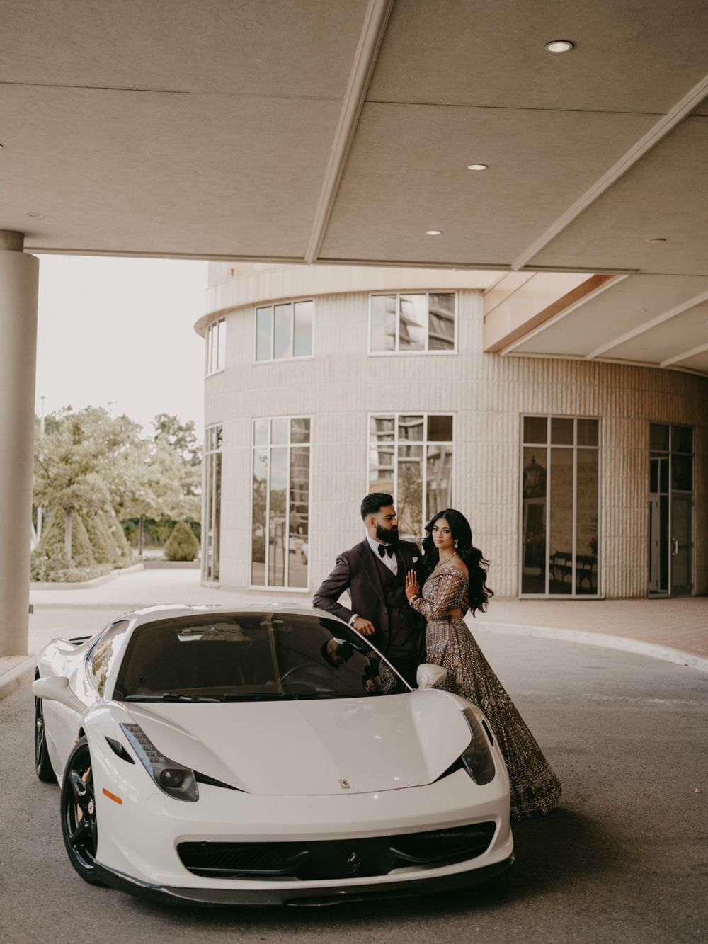 A couple poses with an elegant white Ferrari 458 Italia sports car under a modern building overhang.