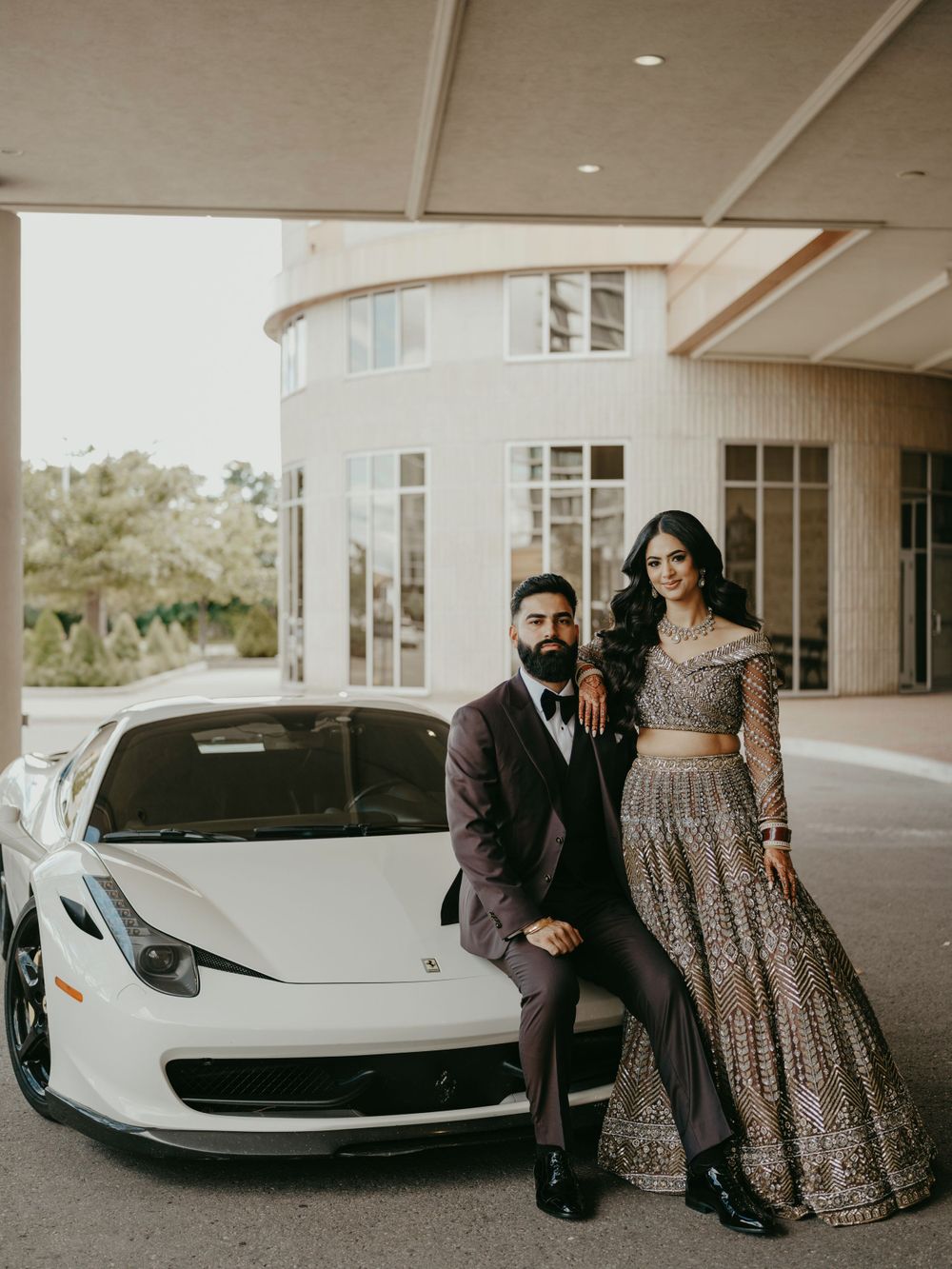 A couple poses with a luxury white sports car outside a modern building wearing formal ethnic attire.