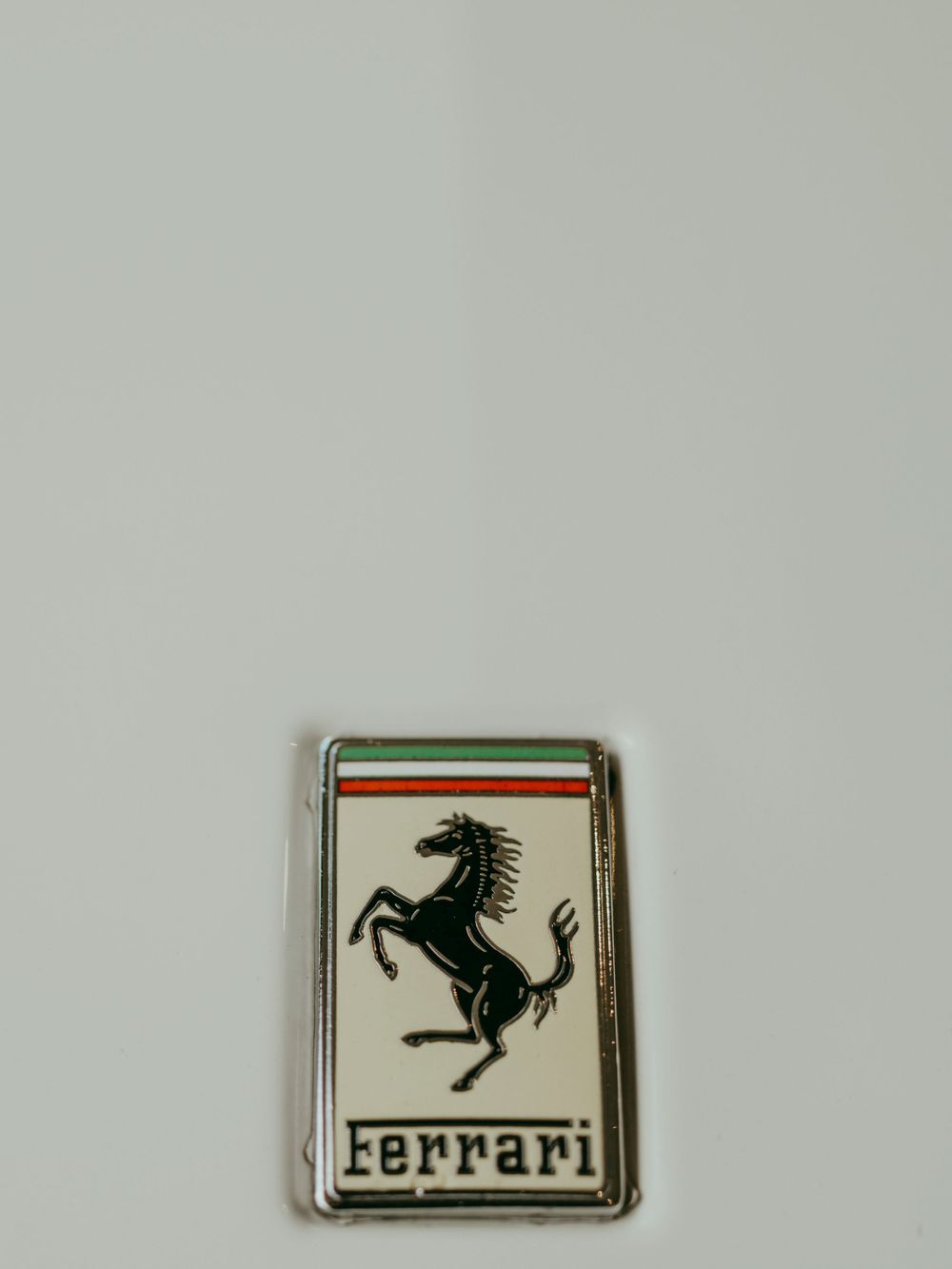 The iconic black Ferrari prancing horse logo emblem against a cream background with Italian flag colors at the top.