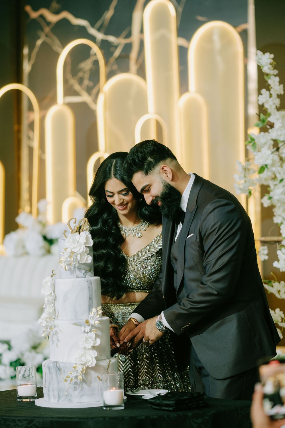 Couple cutting their elegant white wedding cake in front of illuminated art deco backdrop.