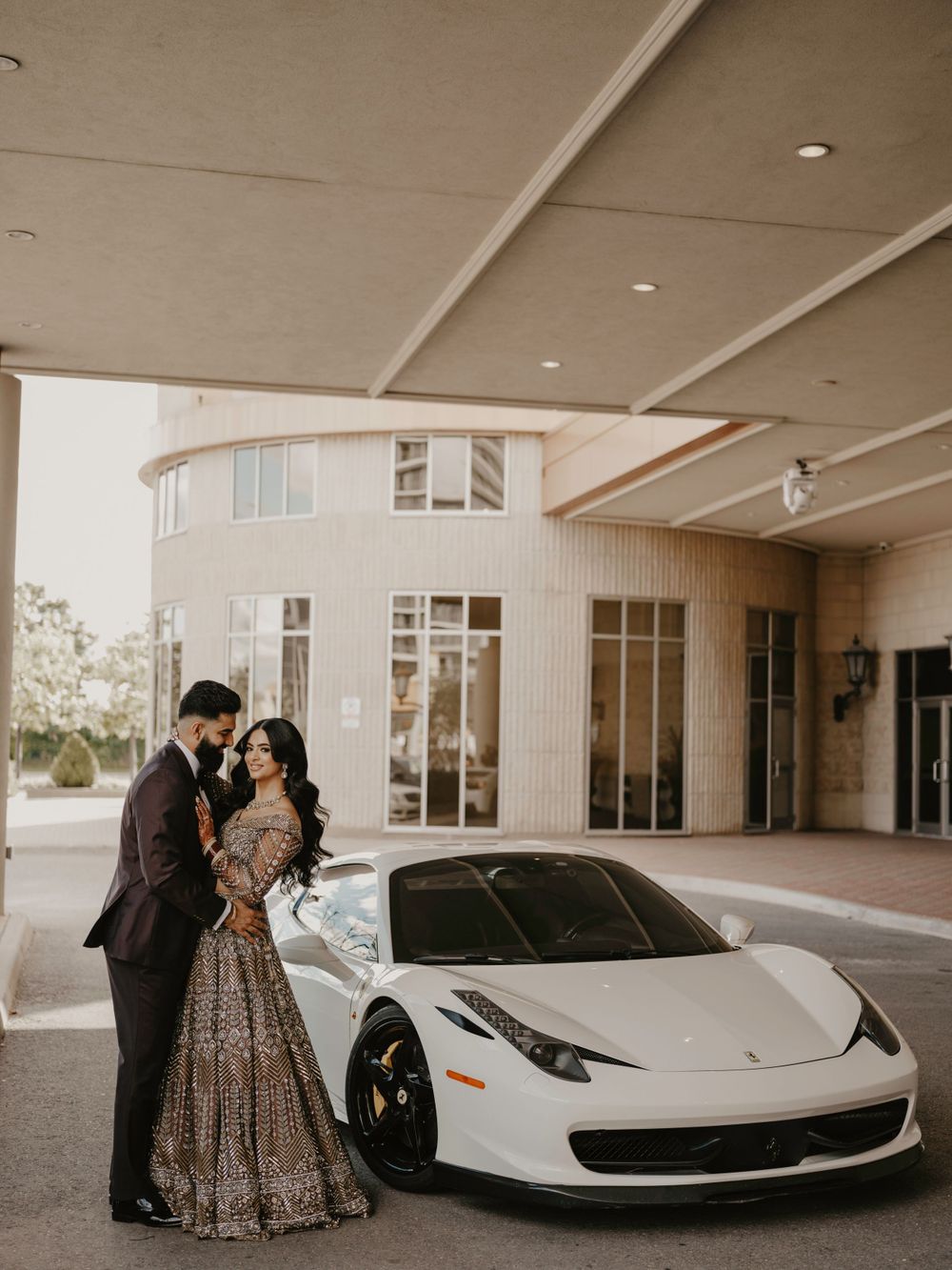 A couple in formal attire poses together next to a white Ferrari 458 Italia outside a modern building entrance.