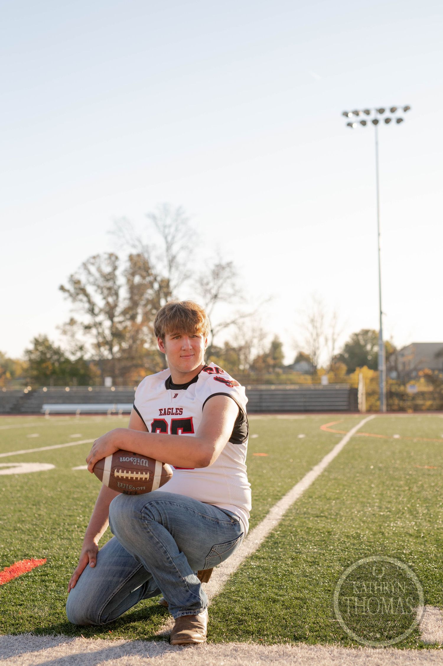Football player kneels on field with ball wearing jersey number 65 at sunset.