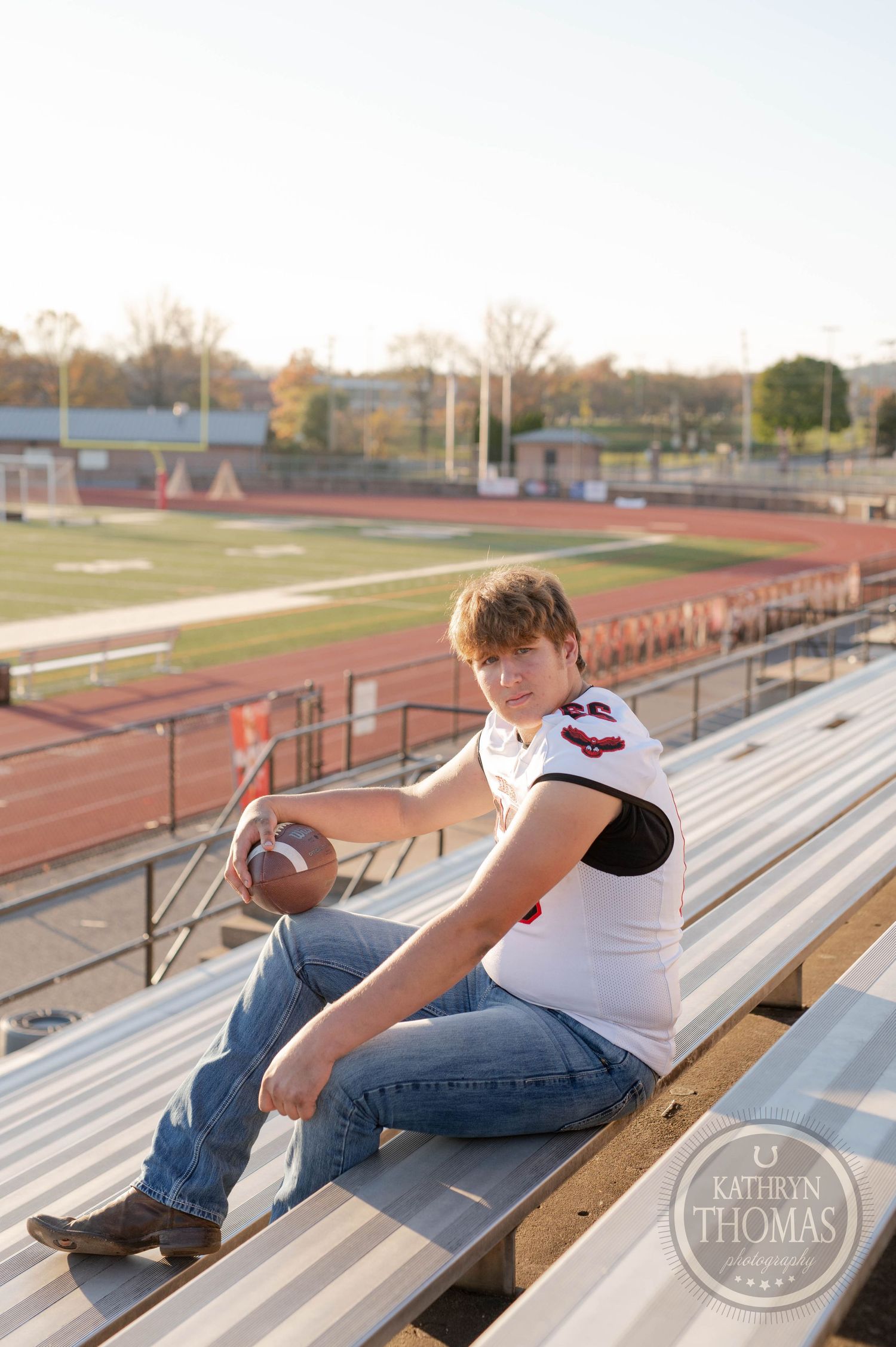 High school athlete in football jersey poses with football on bleachers at sunset with stadium track in background.