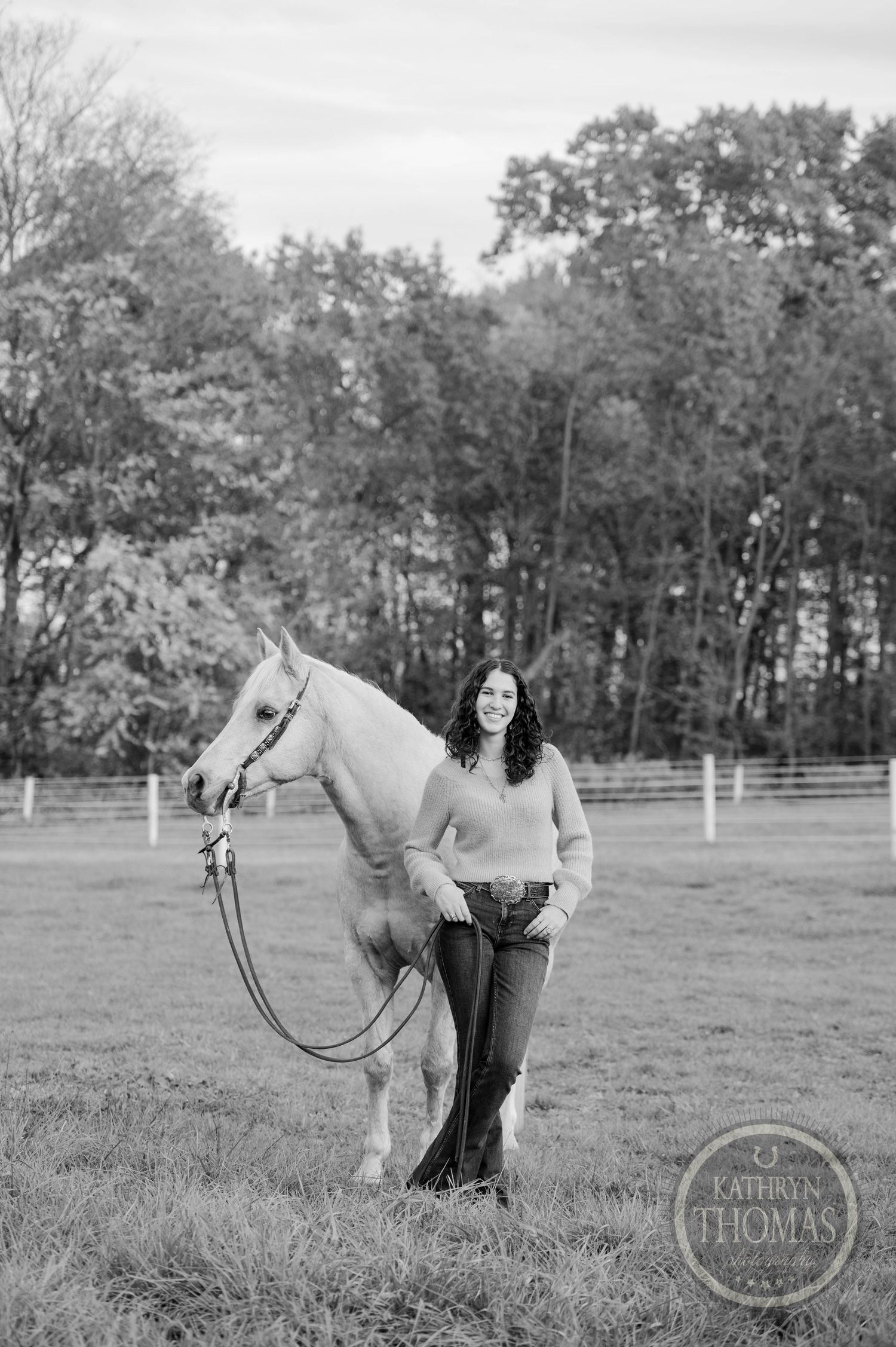 A black and white portrait of a palomino horse and someone in casual attire in a fenced field.