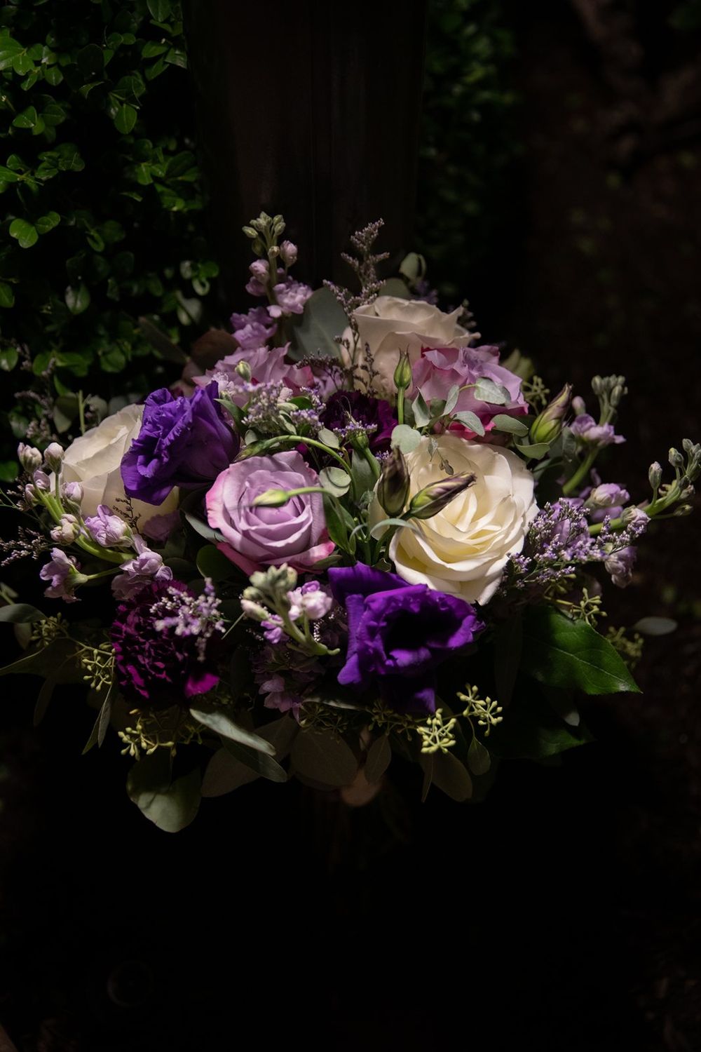 A moody floral arrangement featuring cream and purple roses with delicate lavender accents against a dark background.