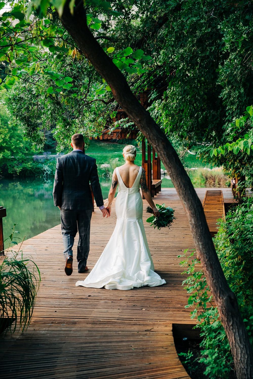 A couple in wedding attire walks along a wooden boardwalk beside a tranquil pond surrounded by lush greenery.