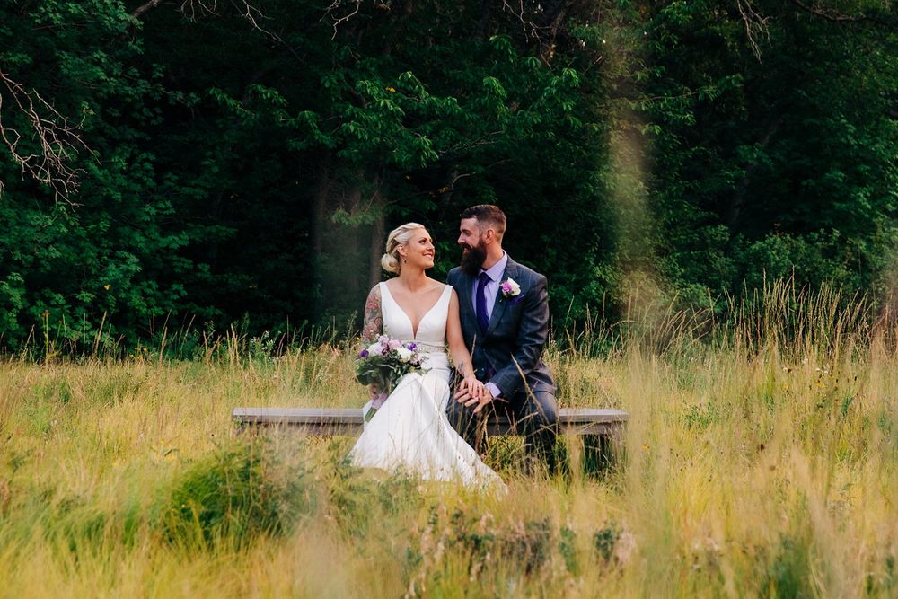 A newlywed couple sits and laughs together on a wooden bench in a scenic meadow surrounded by lush green forest.