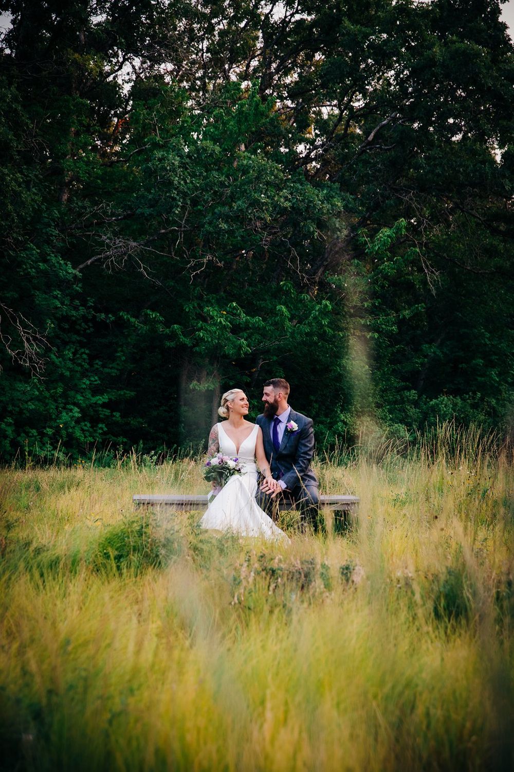 A couple sits together on a rustic bench surrounded by tall grass and green trees in a dreamy outdoor setting at dusk.