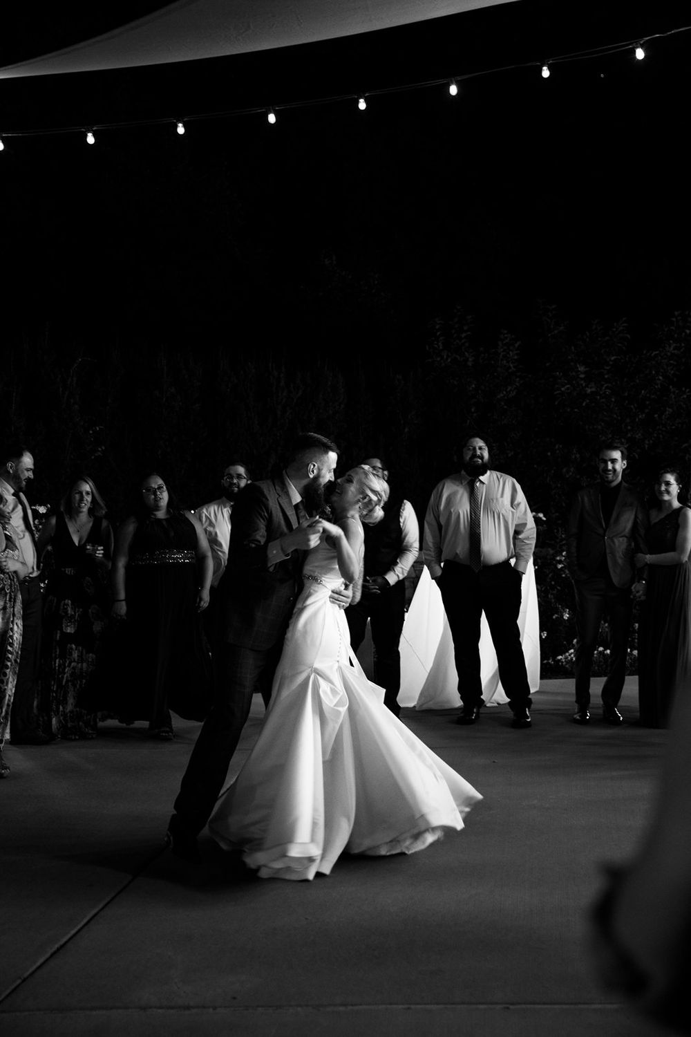 A couple shares an intimate first dance under twinkling lights at their wedding reception while guests look on.