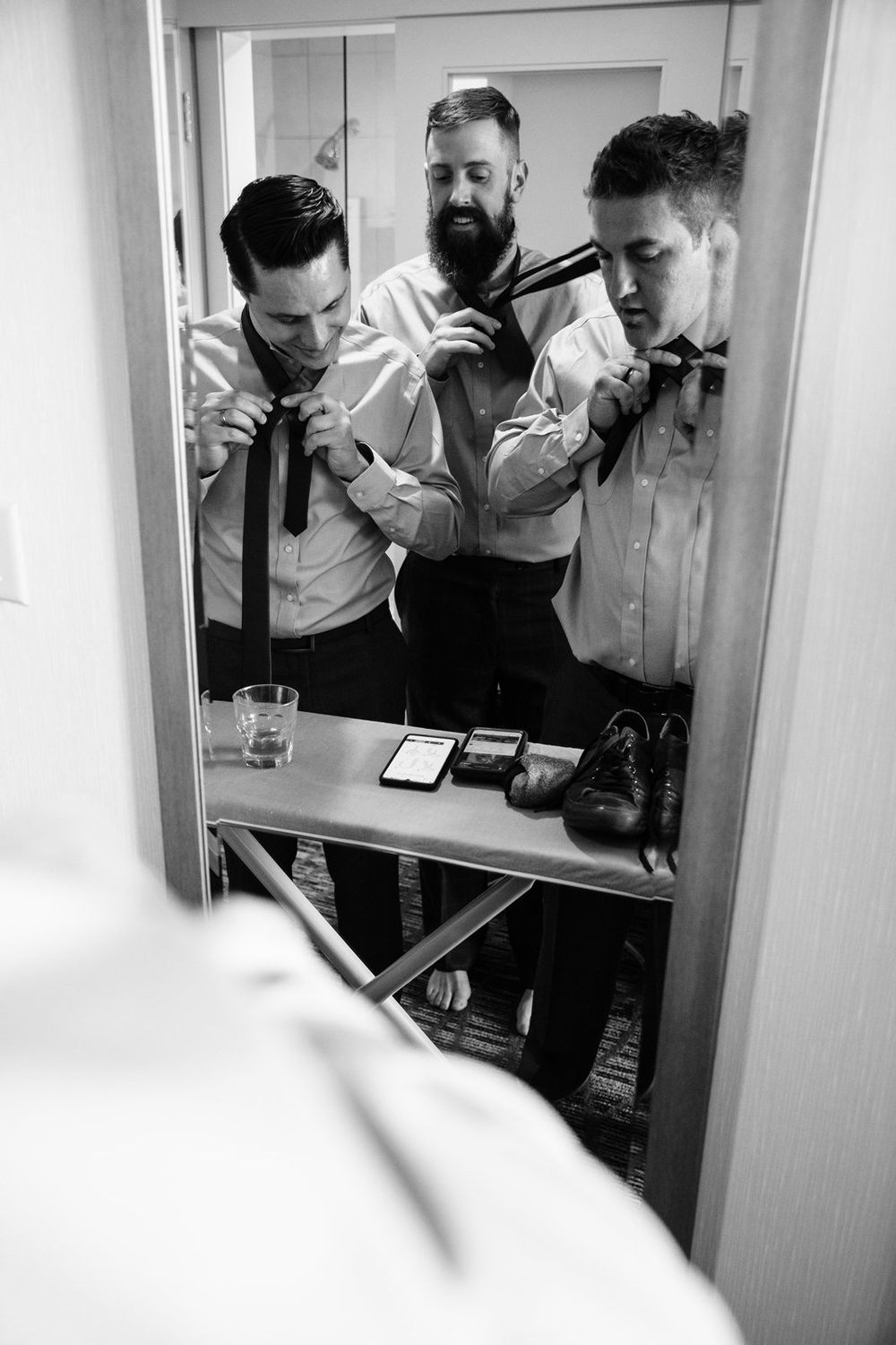 Black and white reflection in mirror shows groomsmen adjusting bow ties while getting ready in formal attire.