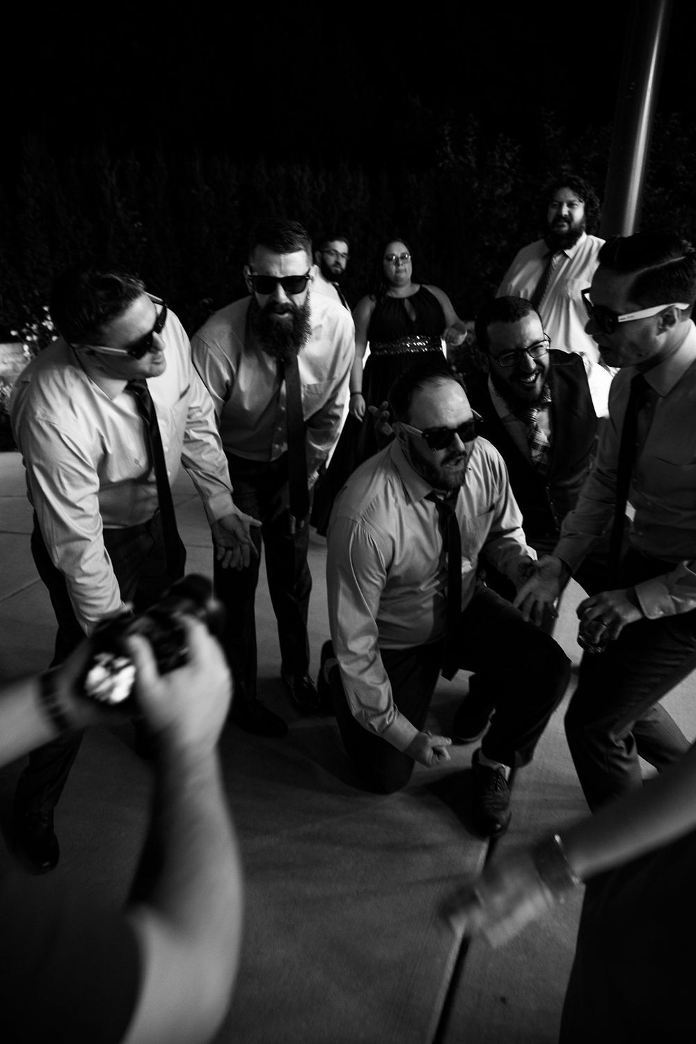 A black and white photo of wedding guests gathered in a circle dancing at a reception celebration.