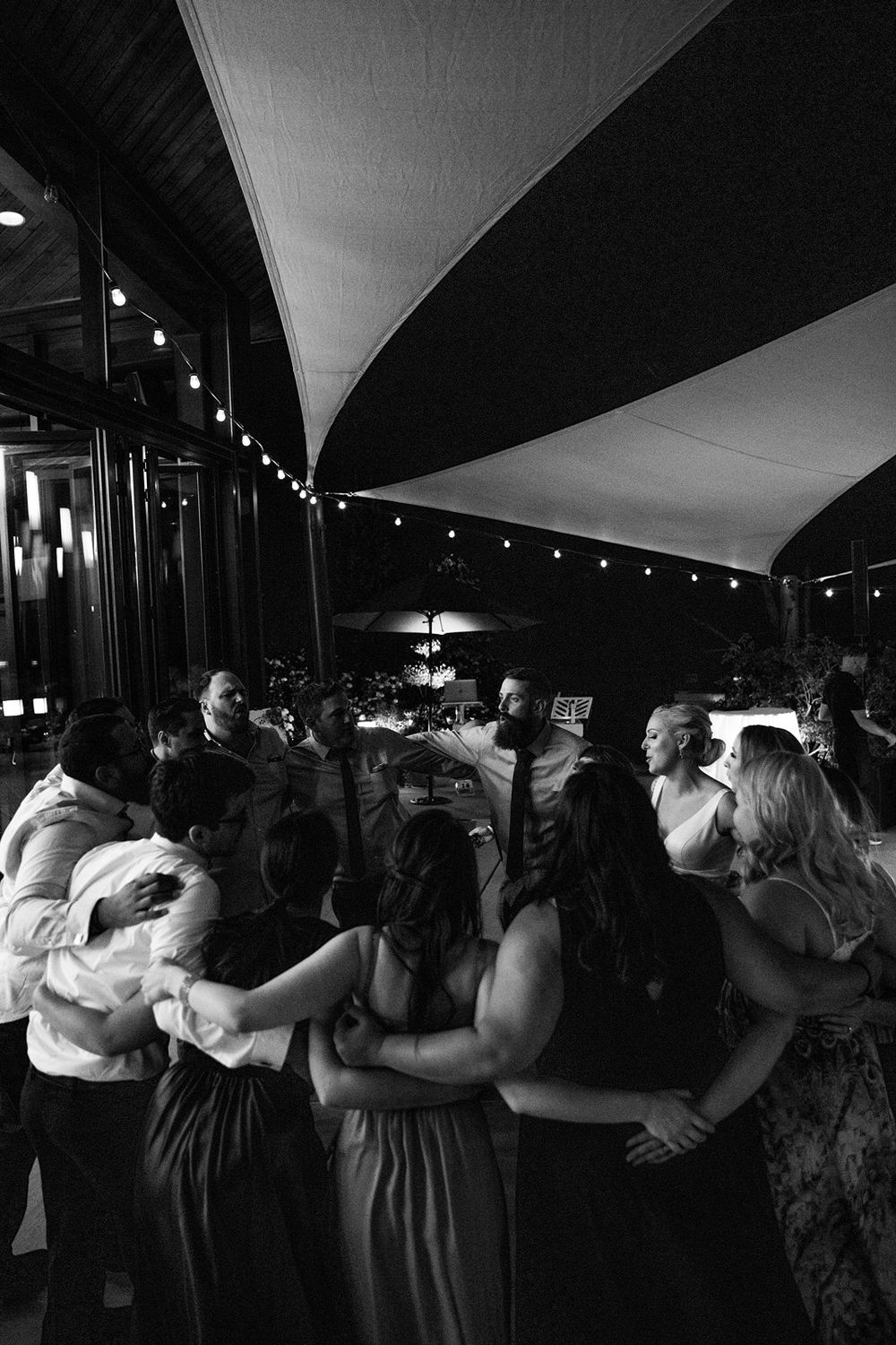 Black and white photo of wedding guests dancing under draped fabric ceiling and string lights.