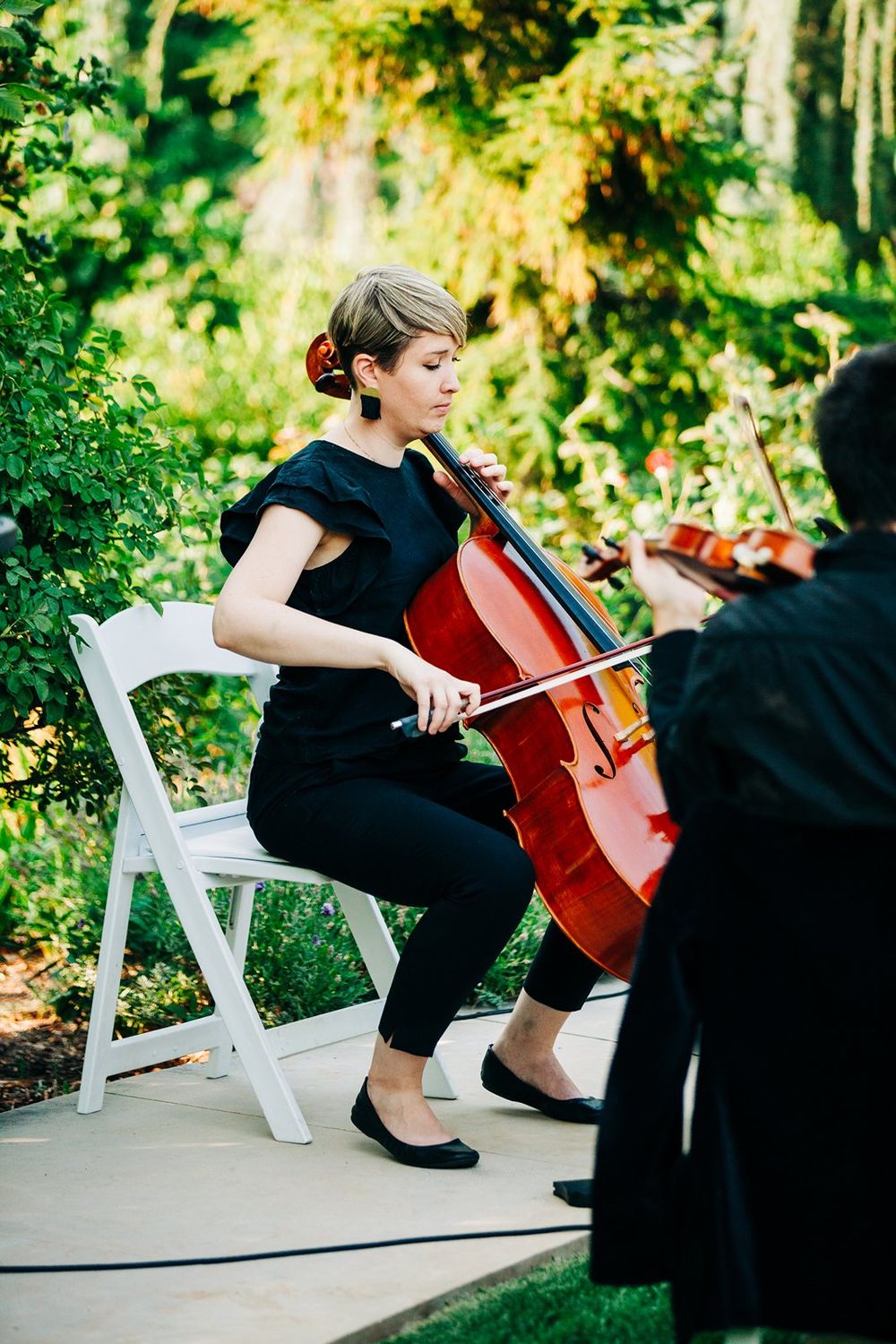 Musician in black outfit plays cello while seated on white folding chair outdoors with lush greenery in background.