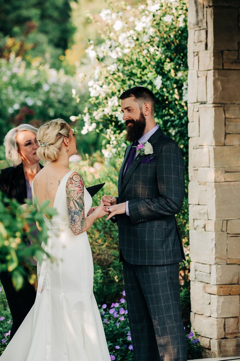 Outdoor wedding ceremony with couple exchanging vows under stone archway surrounded by blooming flowers.