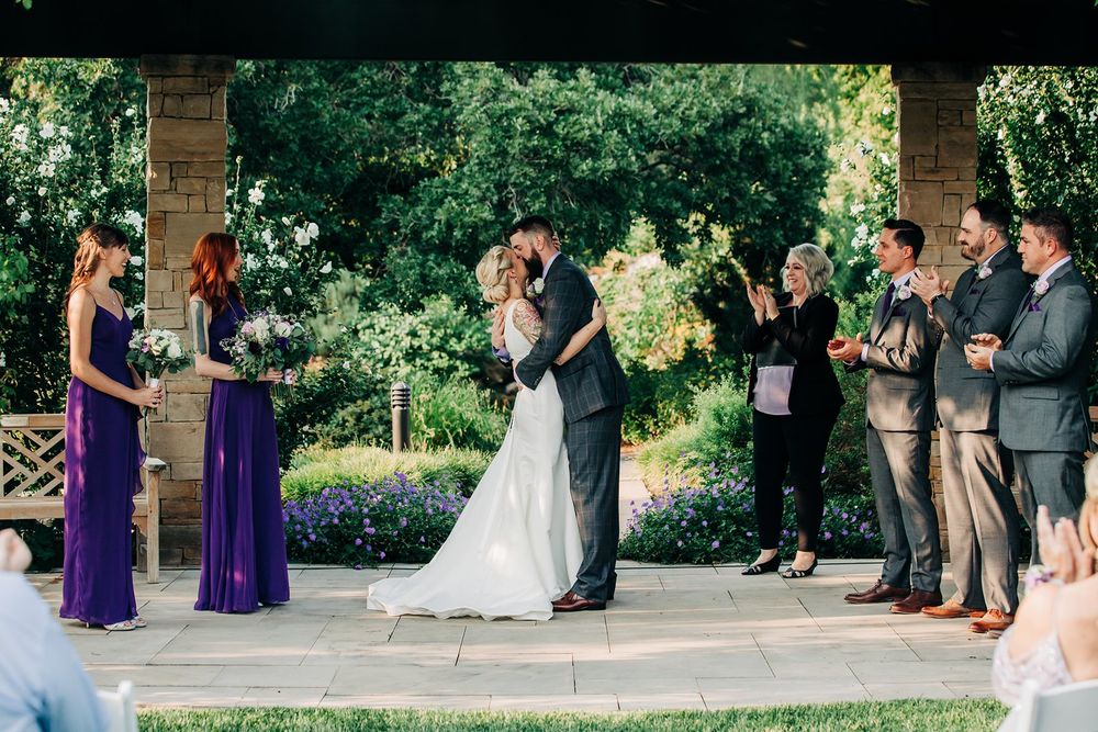 Wedding celebration in a garden pergola with attendants in purple dresses and groomsmen in suits.