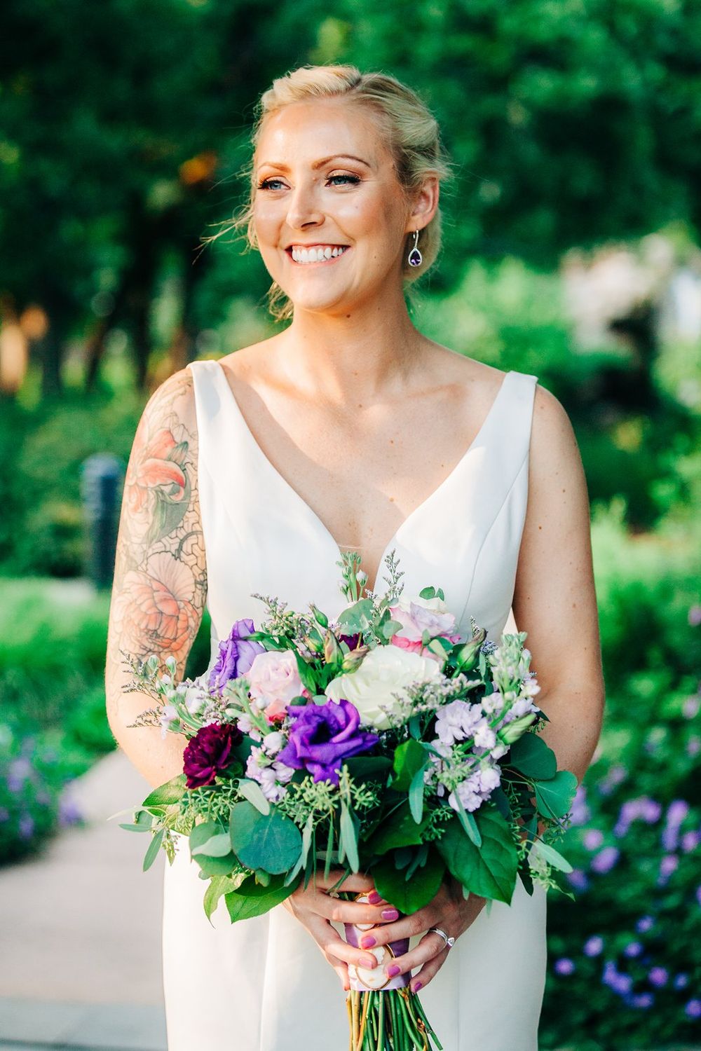 A joyful bride in white dress holds vibrant purple and pink floral bouquet against green garden backdrop.