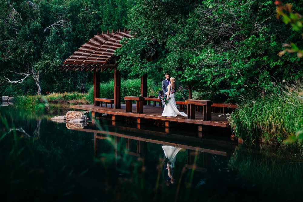 A romantic lakeside scene with a rustic wooden gazebo and dock reflected in calm dark waters surrounded by lush greenery.