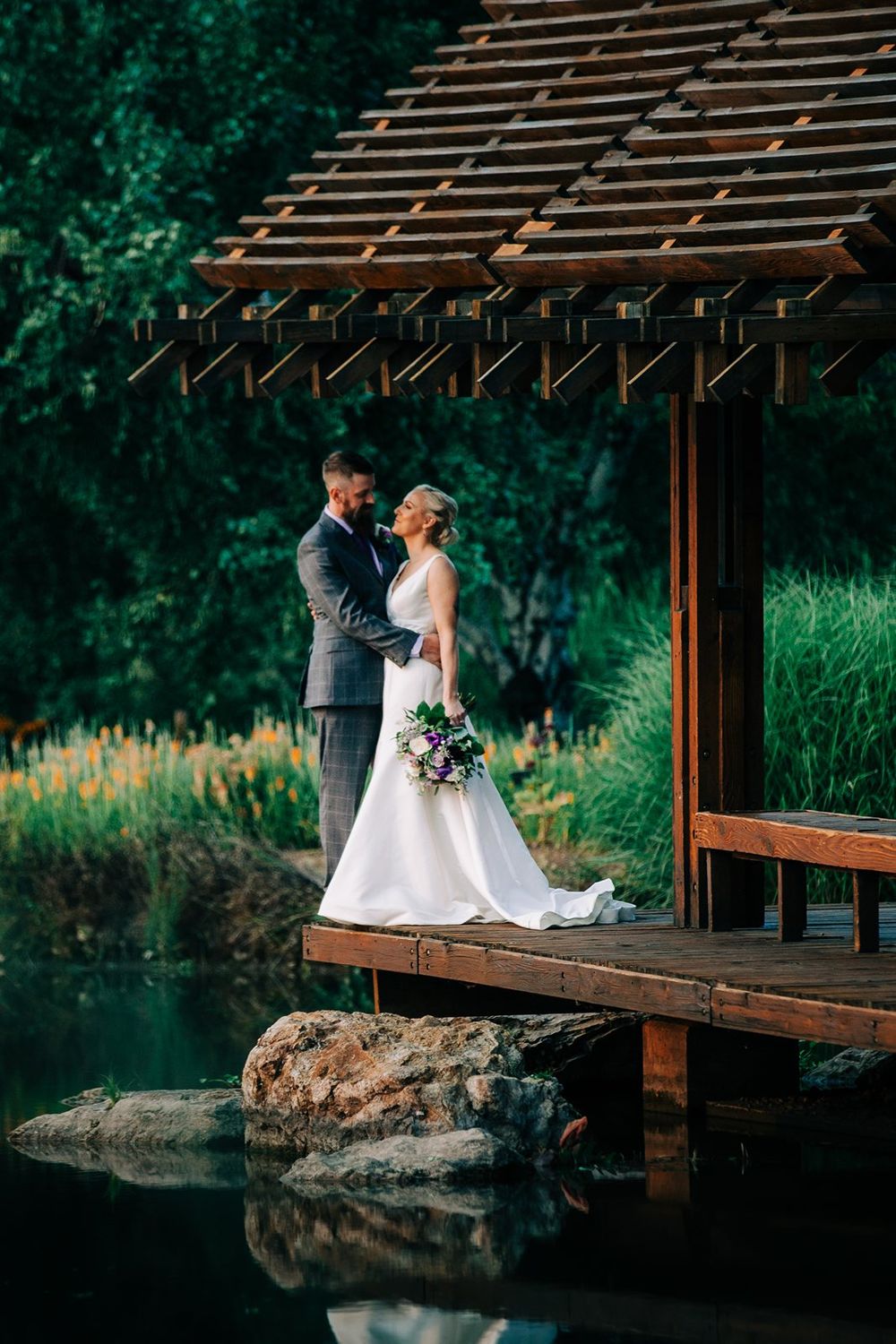 Couple stands together on wooden dock overlooking reflective pond under rustic pergola structure.