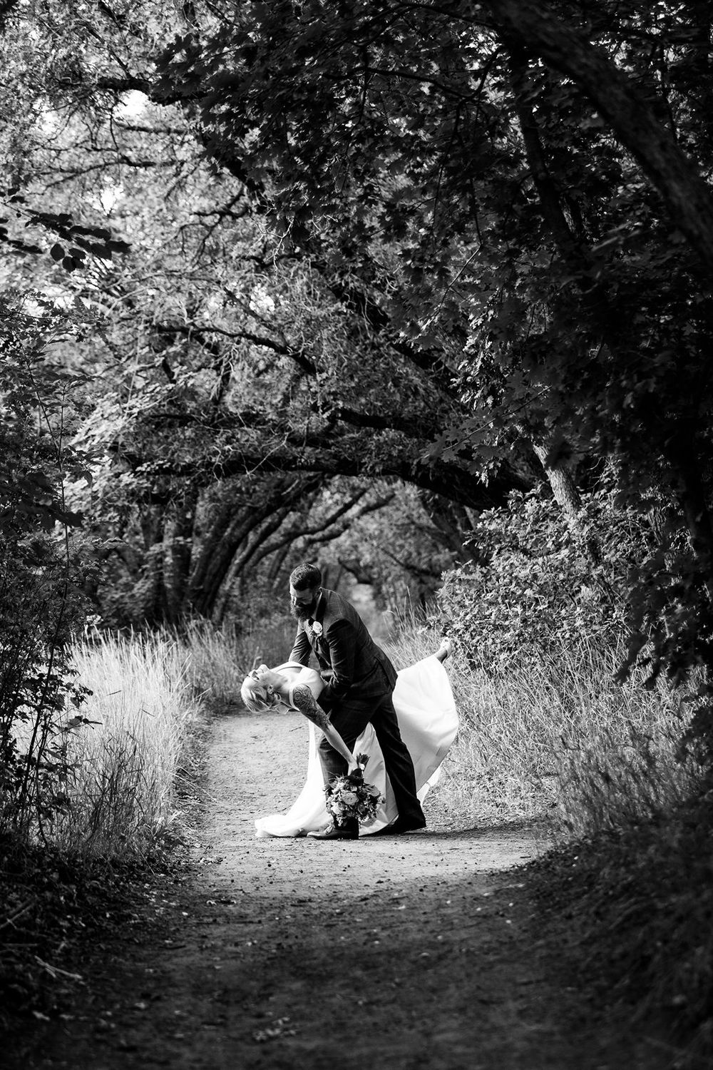 A romantic black and white photograph of a couple dancing on a tree-lined forest path.