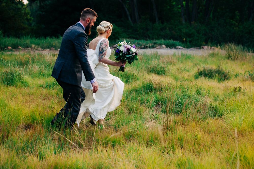 A couple in wedding attire joyfully running together through a lush green meadow at dusk with a bouquet.