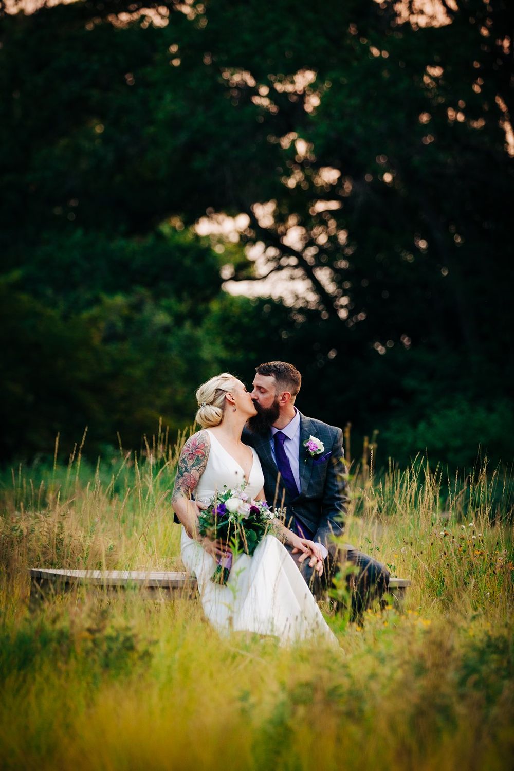 Newlywed couple shares romantic moment while sitting on bench in tall grass field at sunset.