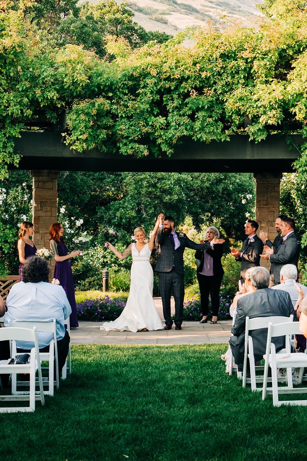 A wedding ceremony under a lush garden pergola with guests seated on white chairs on a manicured lawn.
