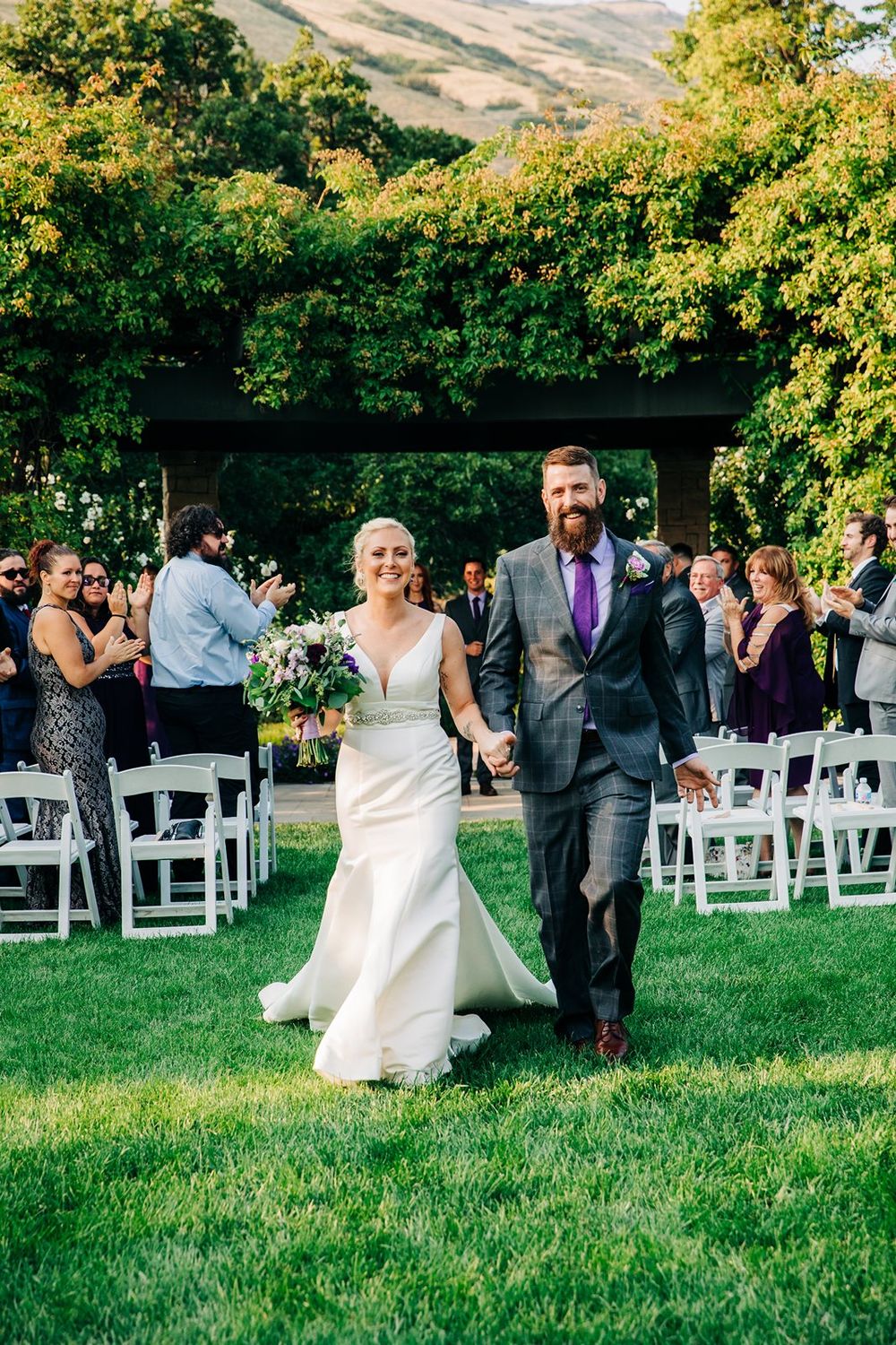 A joyous wedding processional on a lawn with guests seated on white chairs beneath lush green trees.