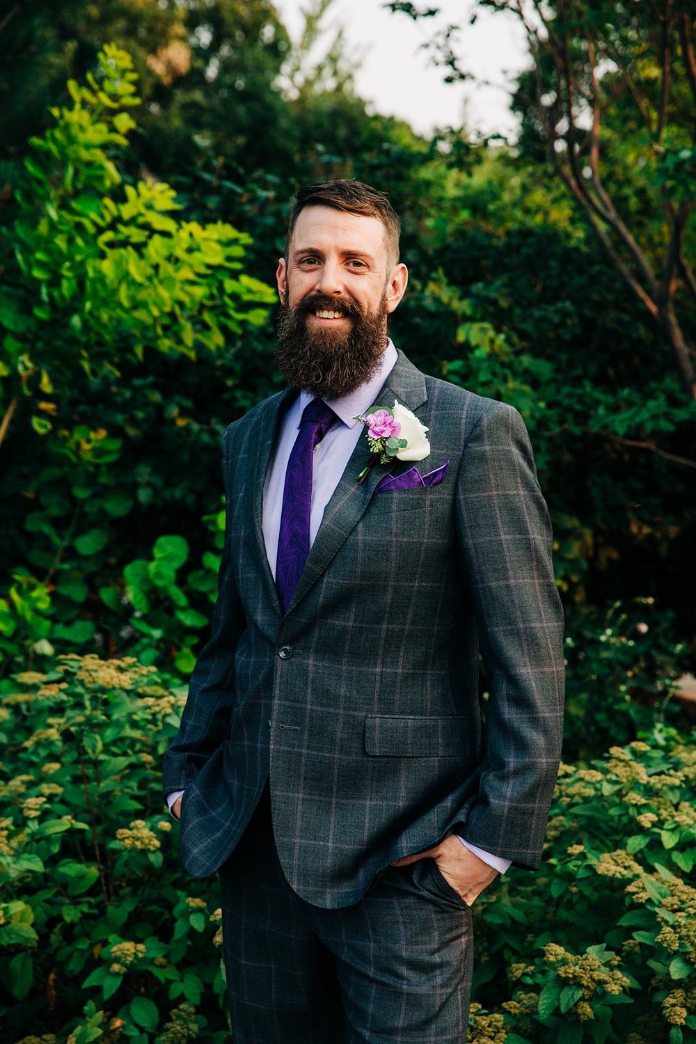 Groom in charcoal windowpane suit with purple tie and boutonniere poses outdoors against lush greenery background.
