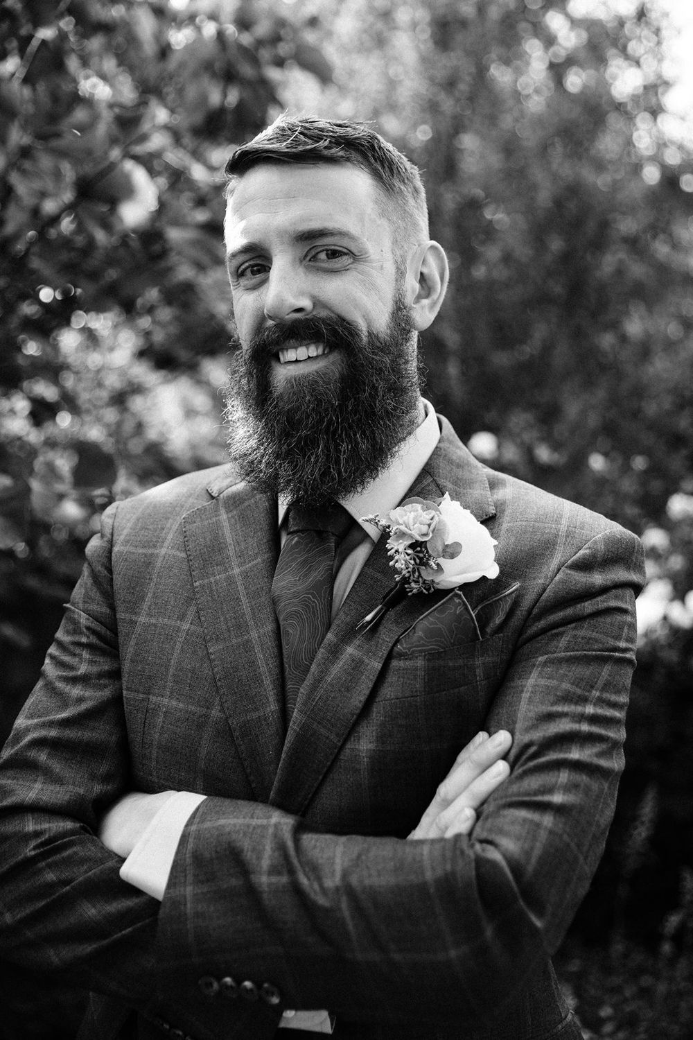 Black and white photo of a groom in a plaid suit with a full beard, flower boutonniere, and crossed arms smiling outdoors.