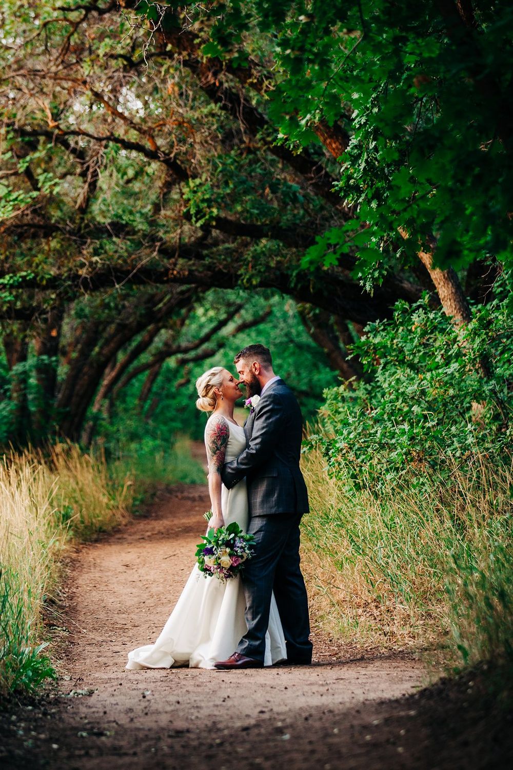 A romantic wedding couple embraces on a tree-lined dirt path surrounded by lush green foliage.