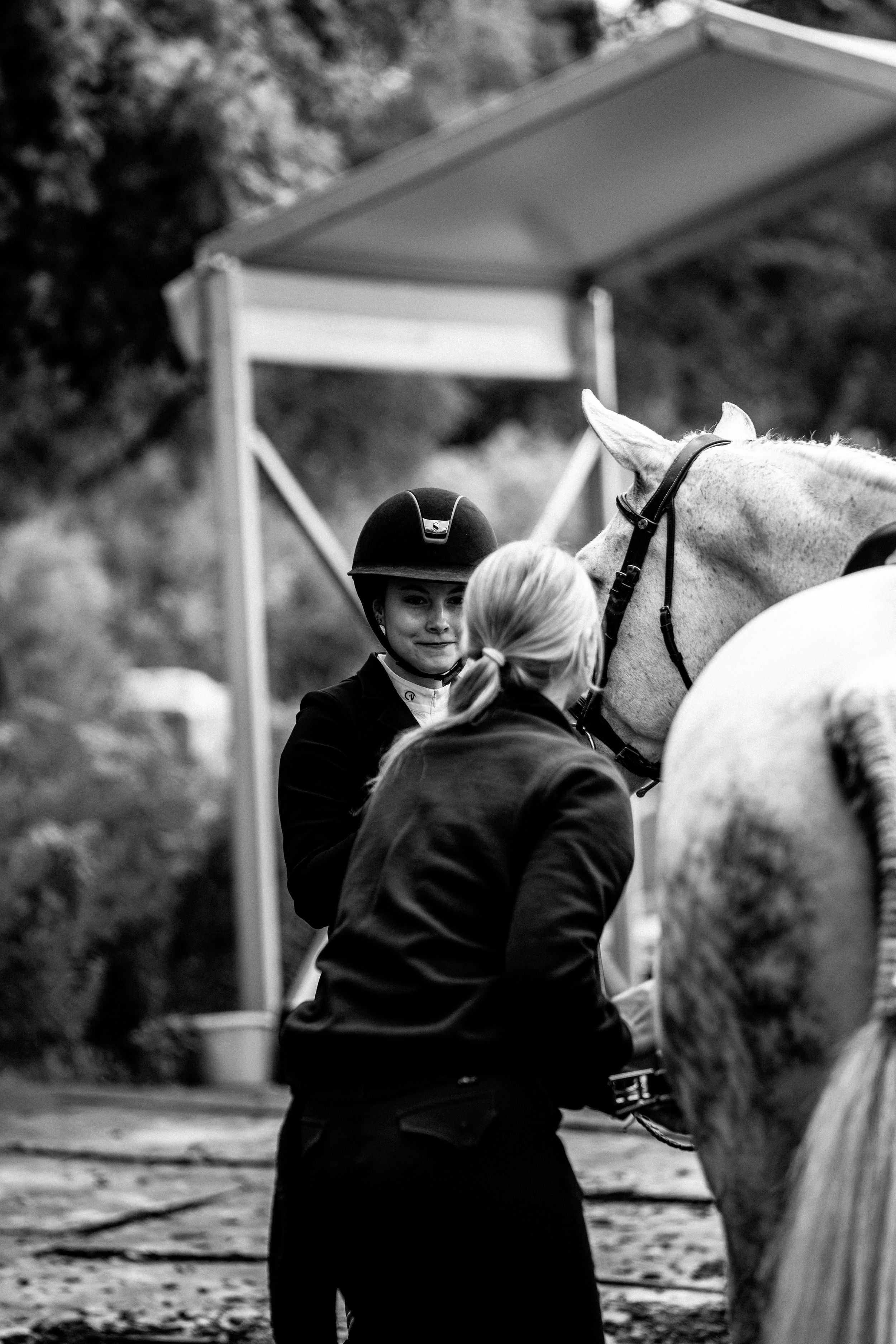 A rider in equestrian attire stands with a white horse during training at an outdoor arena in black and white.