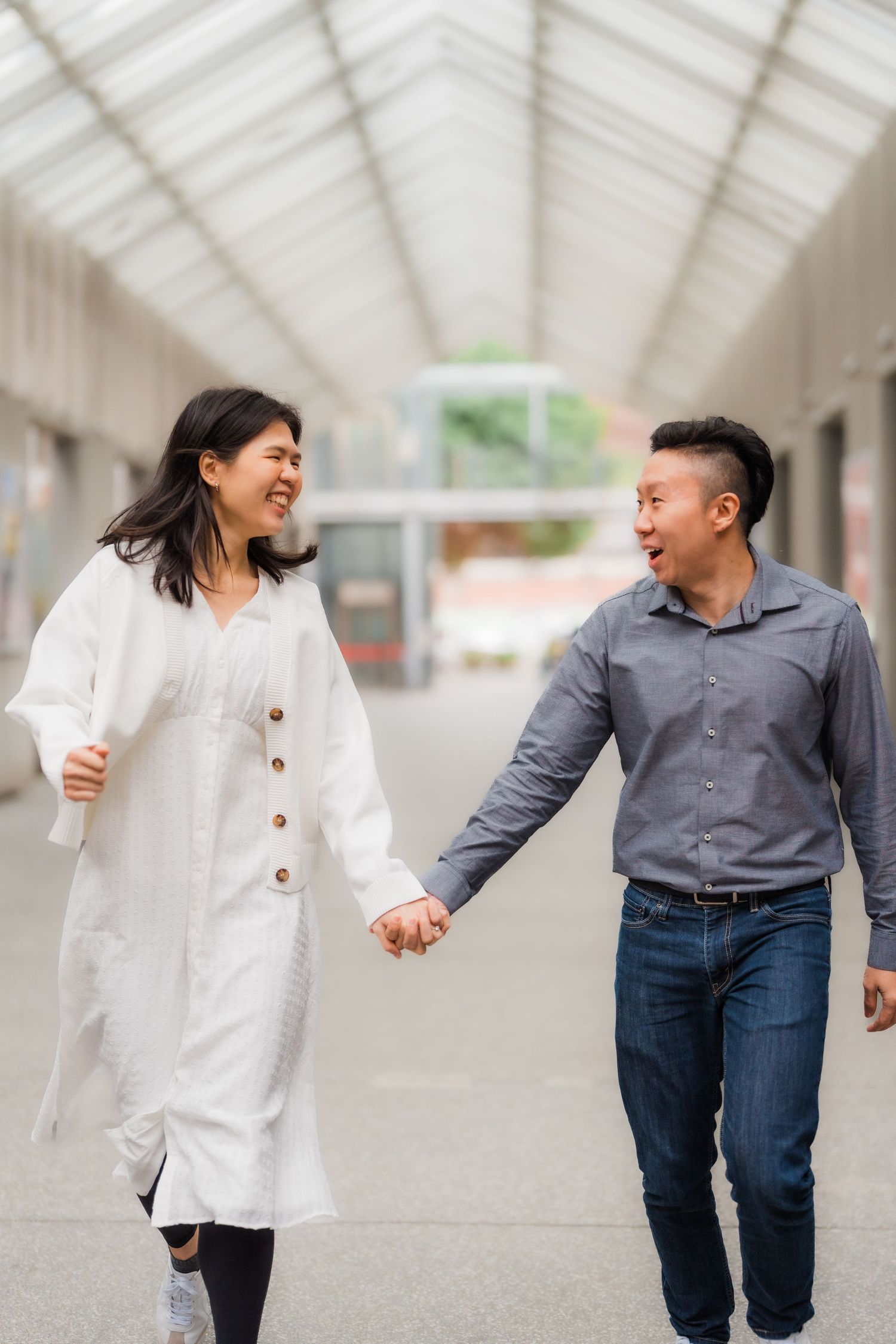 A couple holding hands and laughing while walking together in a bright indoor corridor with glass ceiling.