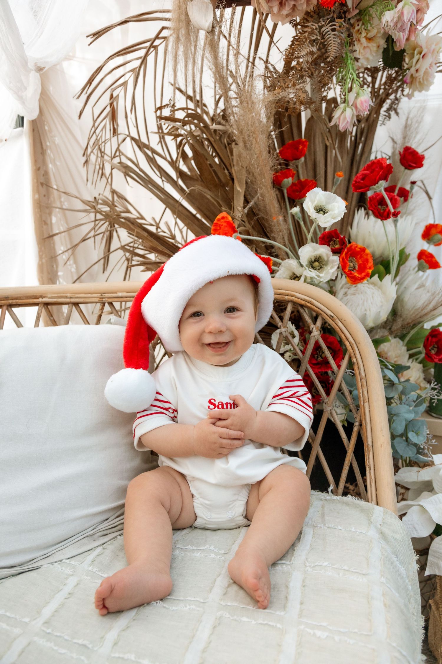 Baby in Christmas attire sitting in wicker chair surrounded by festive floral decorations.