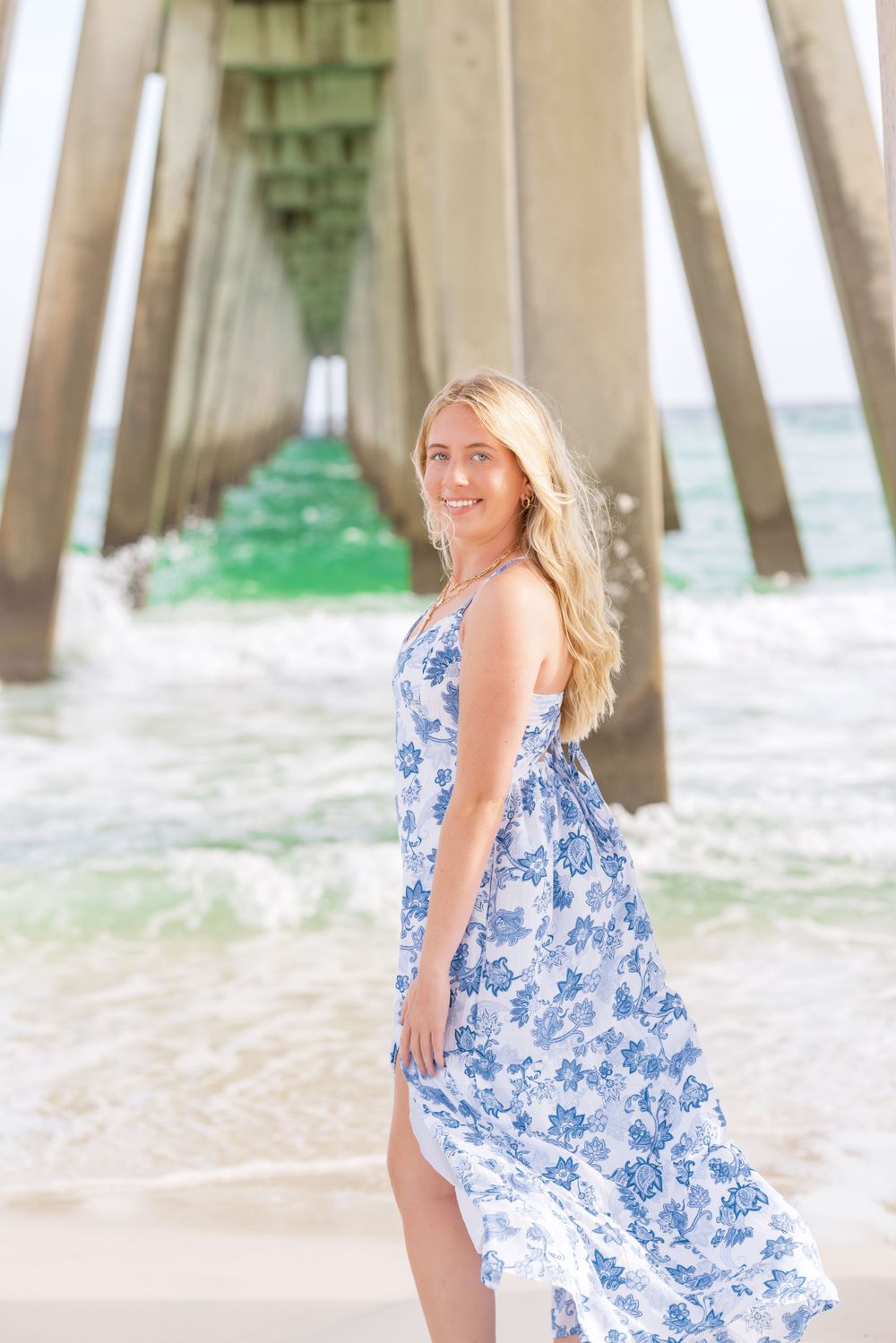 A woman in a flowing blue and white floral dress poses at a beach pier with waves and architectural pillars in the background.