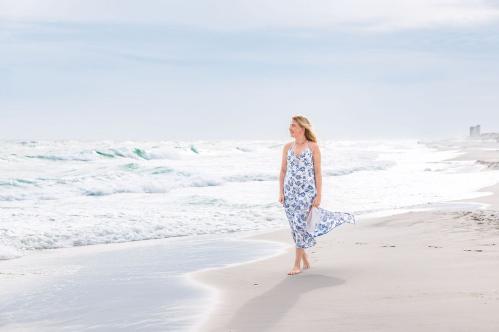 A blue and white floral sundress flows in the ocean breeze as someone walks barefoot along a sandy beach shoreline.