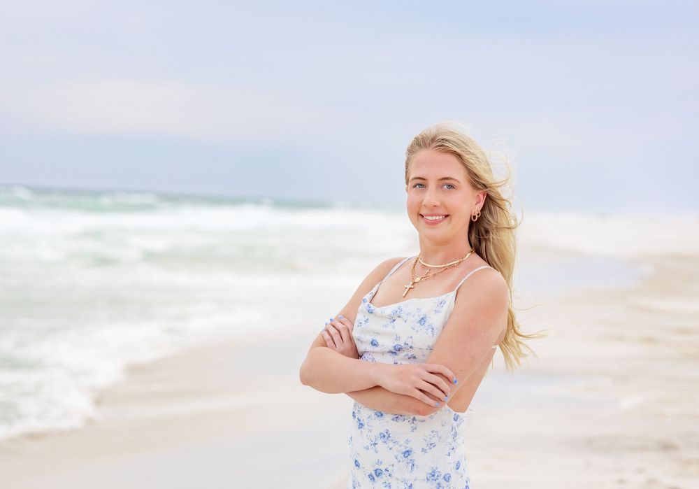A person in a floral blue and white sundress stands with crossed arms on a sunny beach with ocean waves behind them.